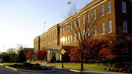 A large brick building with numerous windows, surrounded by autumn trees along a road.