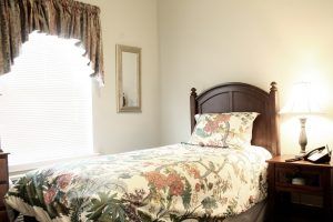 A cozy bedroom with a single bed featuring a floral bedspread, accompanied by a window with curtains and a wall mirror.