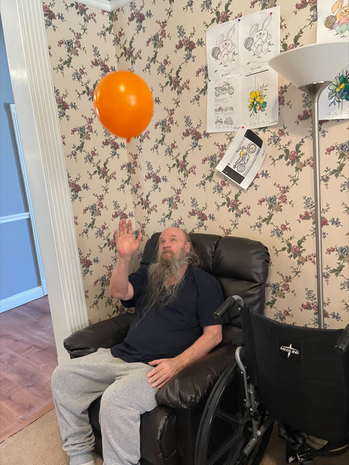 A man seated on a leather chair is playfully tossing an orange balloon indoors.