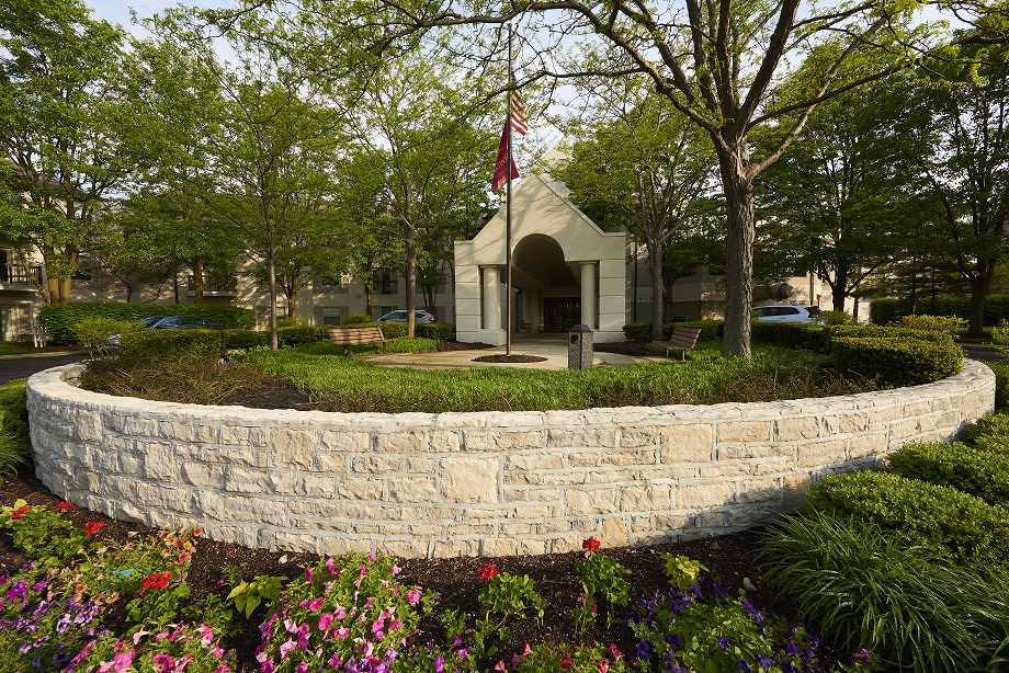 A scenic view of a small building surrounded by trees, flowers, and a stone wall, with a flag flying in the foreground.