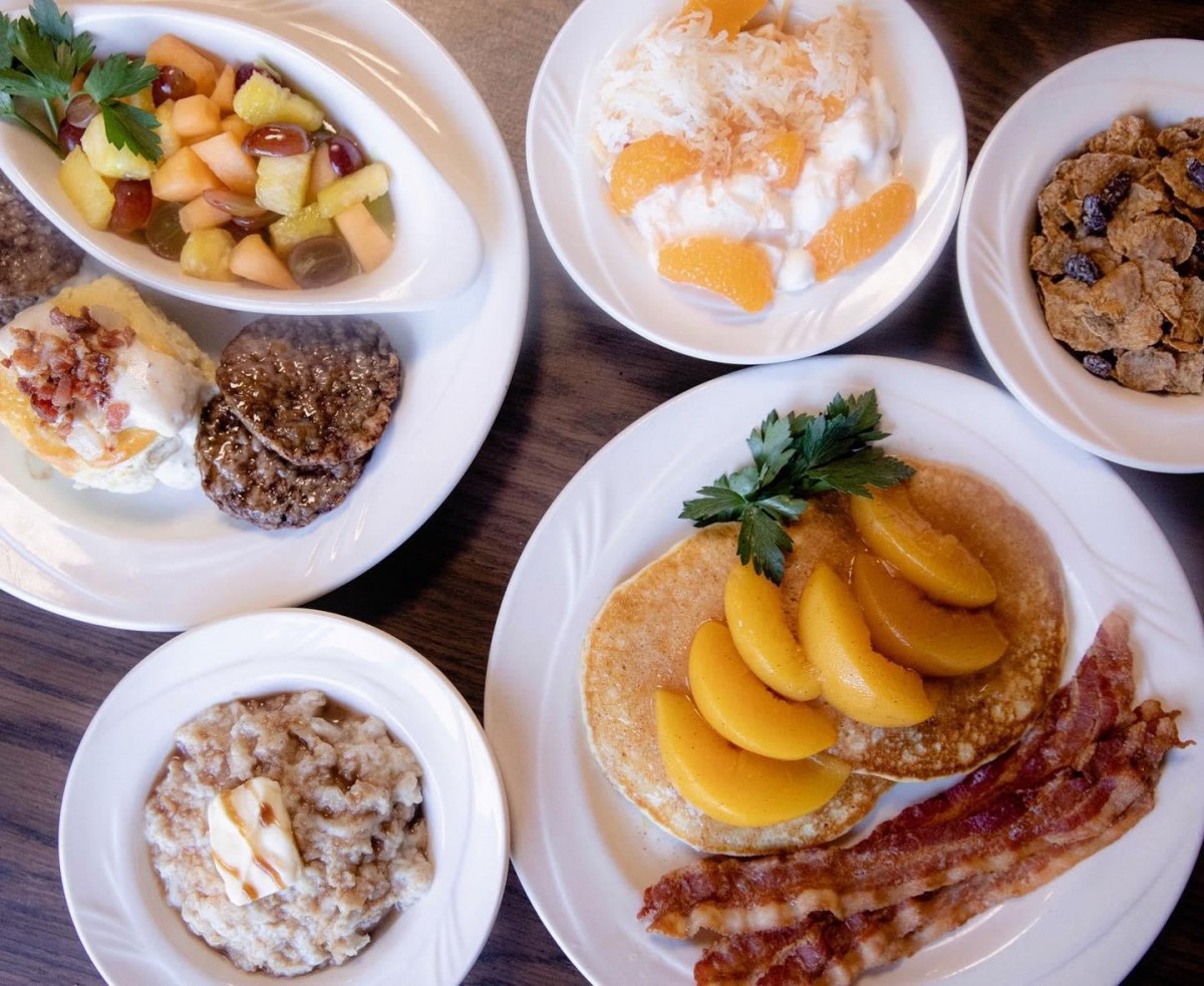 A hearty breakfast spread featuring fruit-topped pancakes, eggs, bacon, and cereal.
