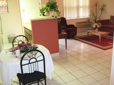 A cozy living and dining area with tiled flooring, featuring a small dining table, chairs, armchair, and potted plants.