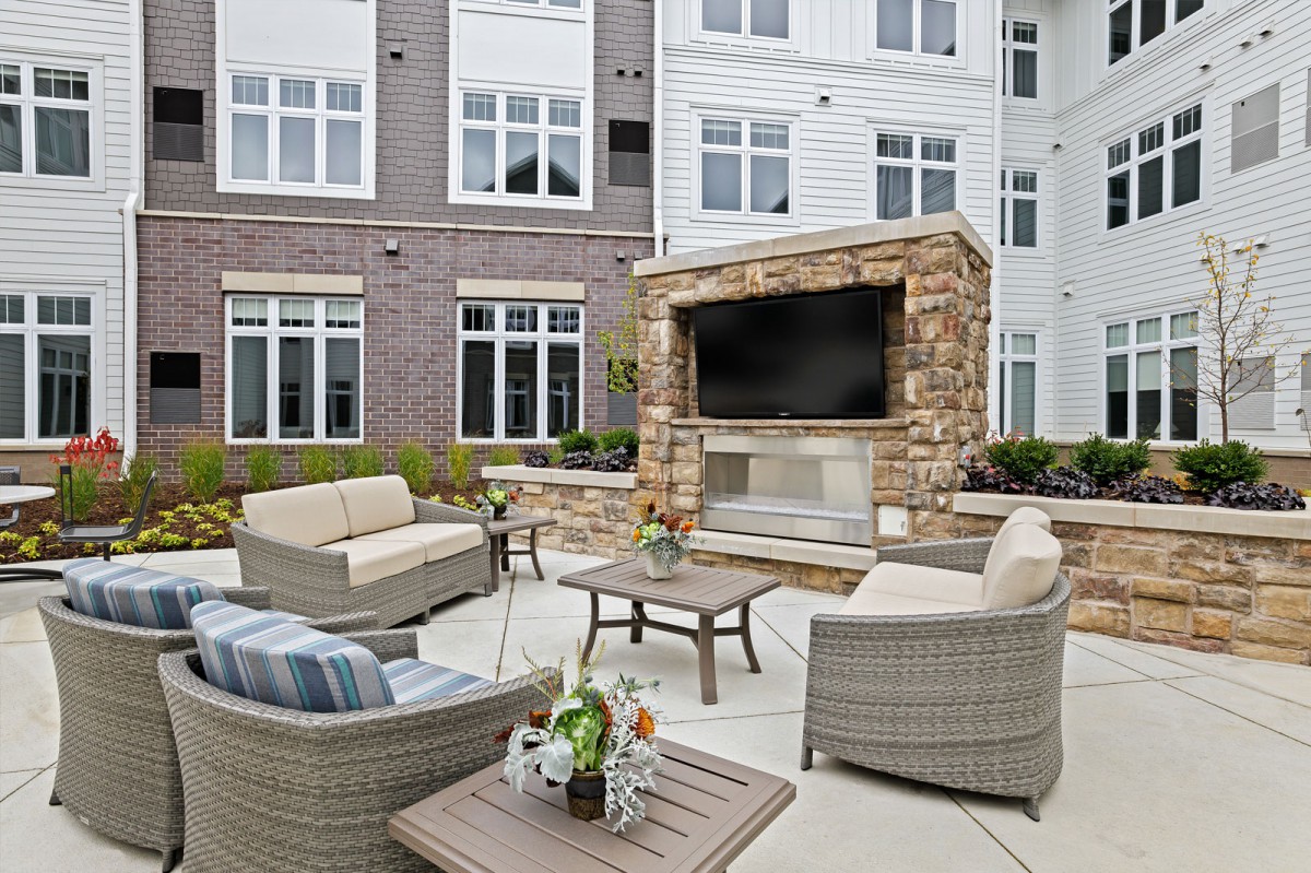 A modern outdoor seating area with a stone fireplace and television, set in front of an apartment building.