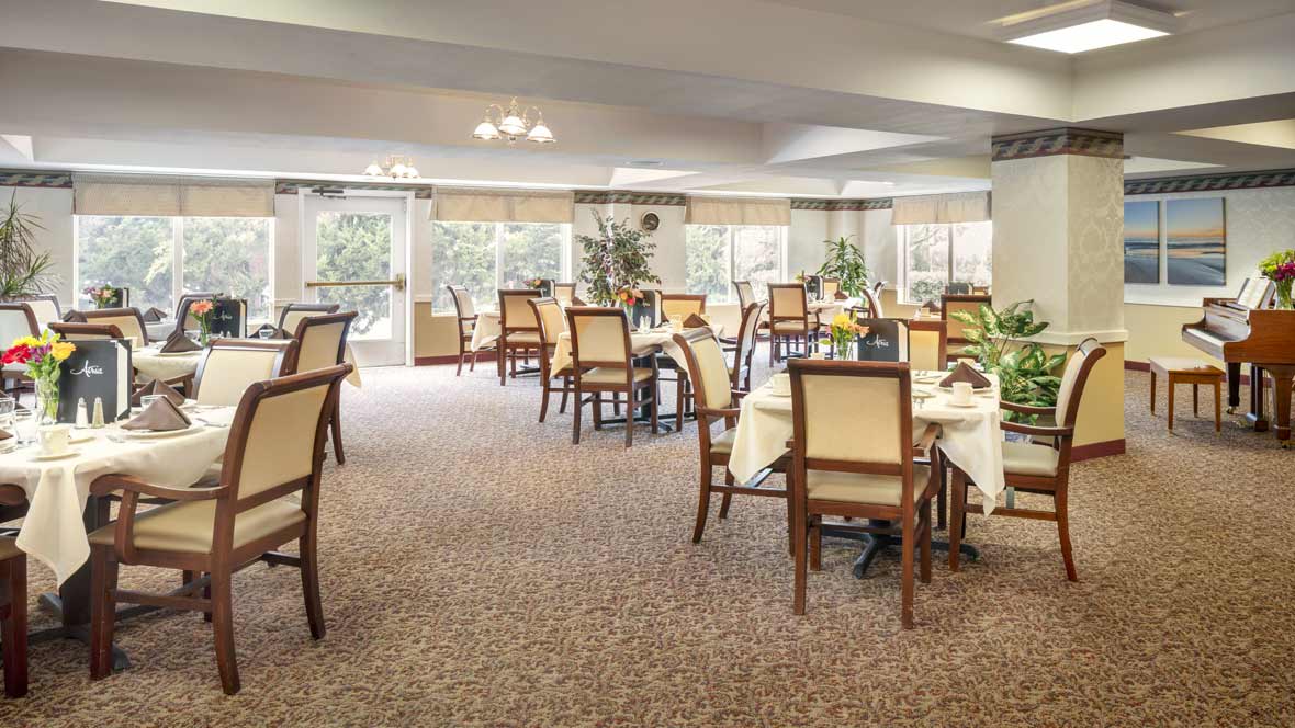 A spacious and elegantly decorated dining room with several tables and chairs, featuring large windows and a carpeted floor.