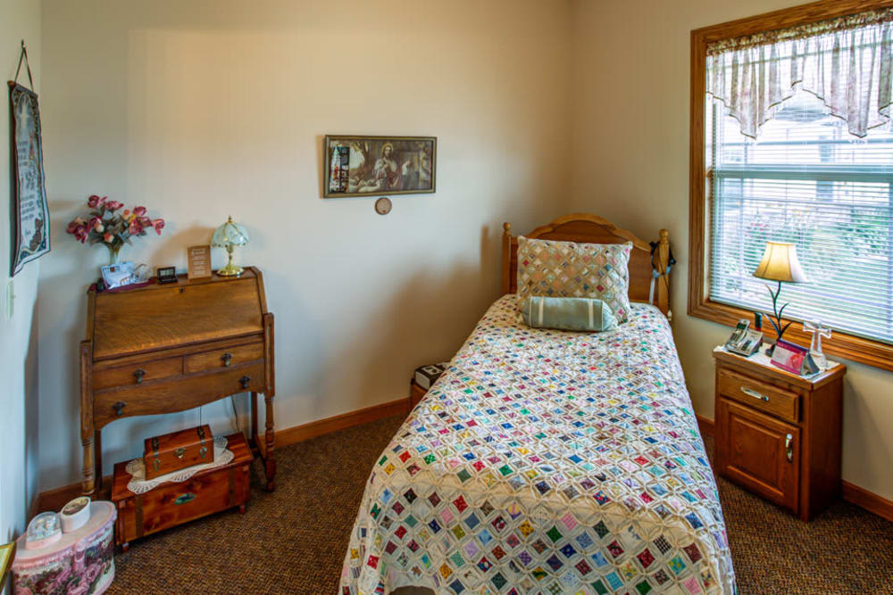 A cozy bedroom with a single bed featuring a colorful quilt, wooden desk, and window.