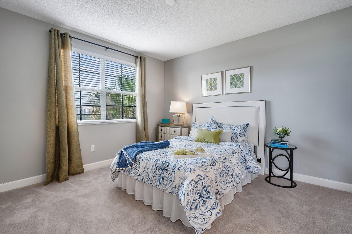A well-lit bedroom with a patterned bedspread, a bedside lamp, and framed artwork on the wall.