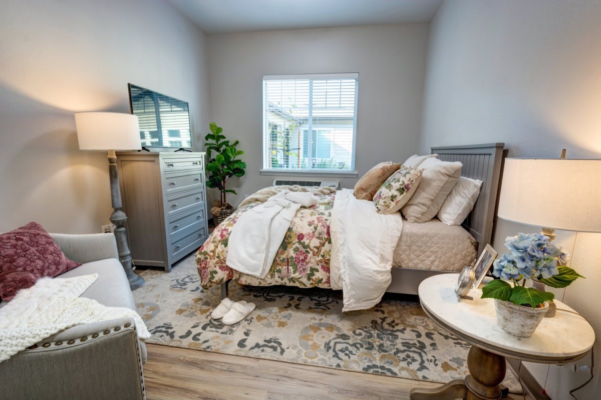 A cozy, well-decorated bedroom featuring a floral bedspread, stylish dresser, and natural light from the window.