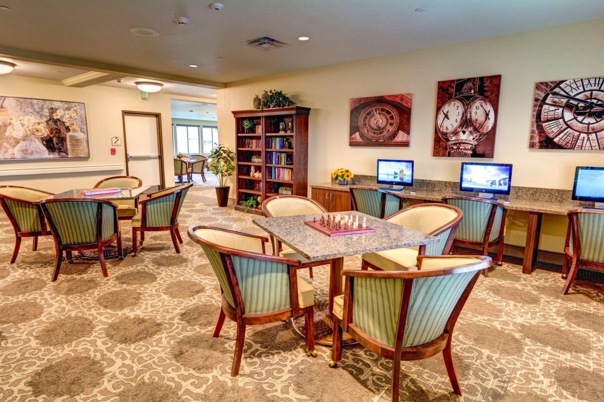A cozy library room with tables, chairs, computers, and a bookshelf.
