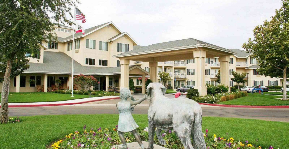 A large building with a covered entrance, featuring a statue of a girl and a horse in the foreground, surrounded by a garden.