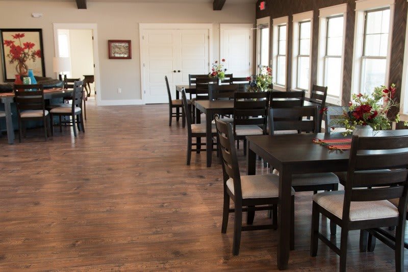 A spacious dining area with multiple dark wooden tables and chairs, featuring large windows and a wooden floor.