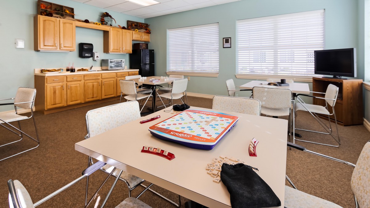 A community game room with a Scrabble board set up on a table, surrounded by chairs and featuring a small kitchenette area.