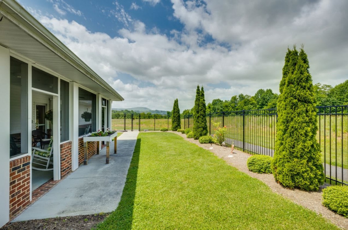 A spacious backyard with a green lawn and a metal fence, surrounded by trees under a partly cloudy sky.