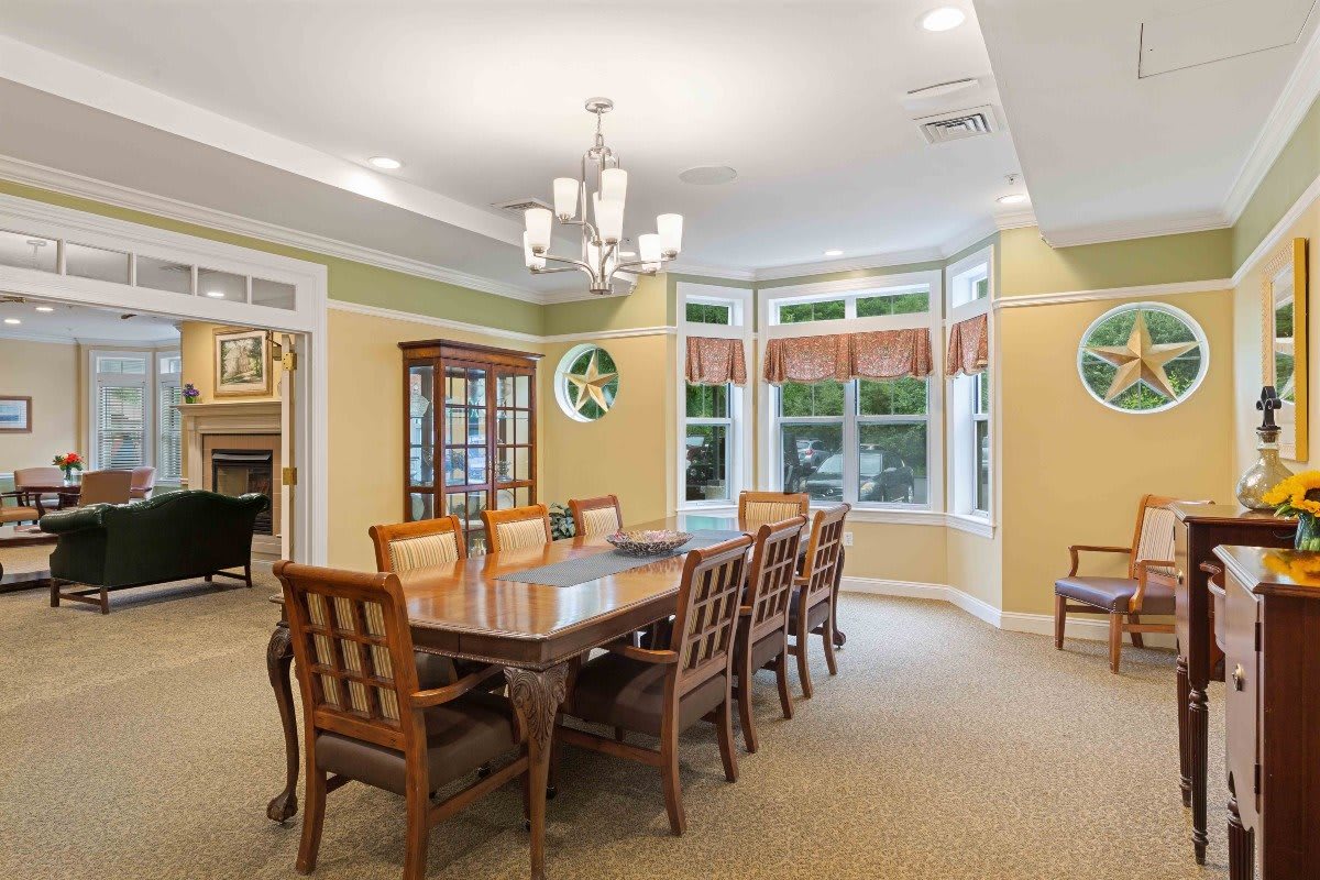A well-lit dining room featuring a long wooden table with matching chairs, a chandelier overhead, large windows with curtains, and a wooden cabinet.
