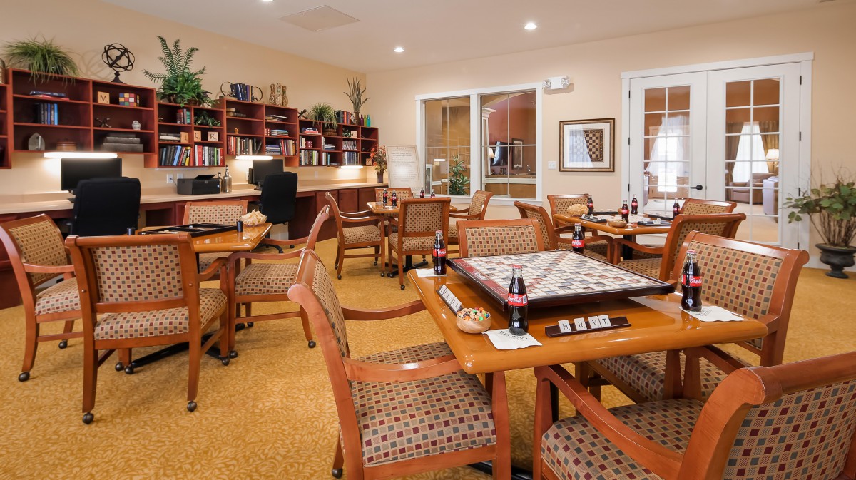 A cozy game room with a Scrabble board set up on a wooden table surrounded by comfortable chairs and bookshelves in the background.