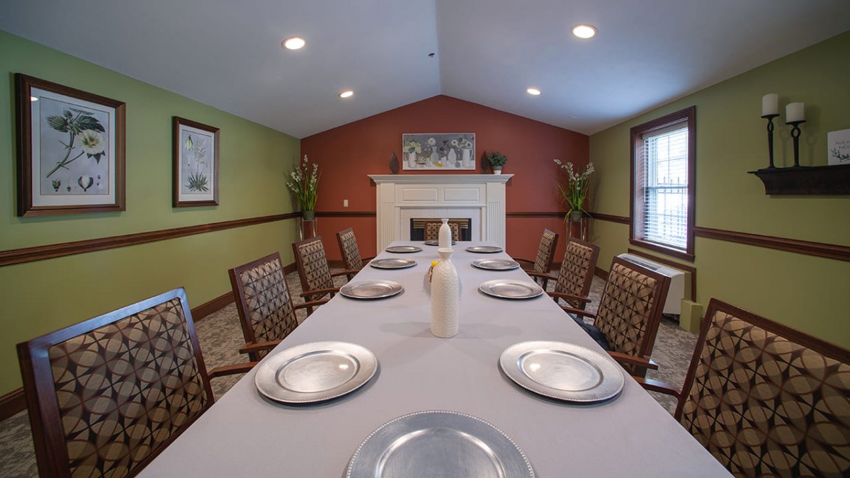 A dining room with a long table set for a meal, featuring plates and chairs, and a fireplace in the background.