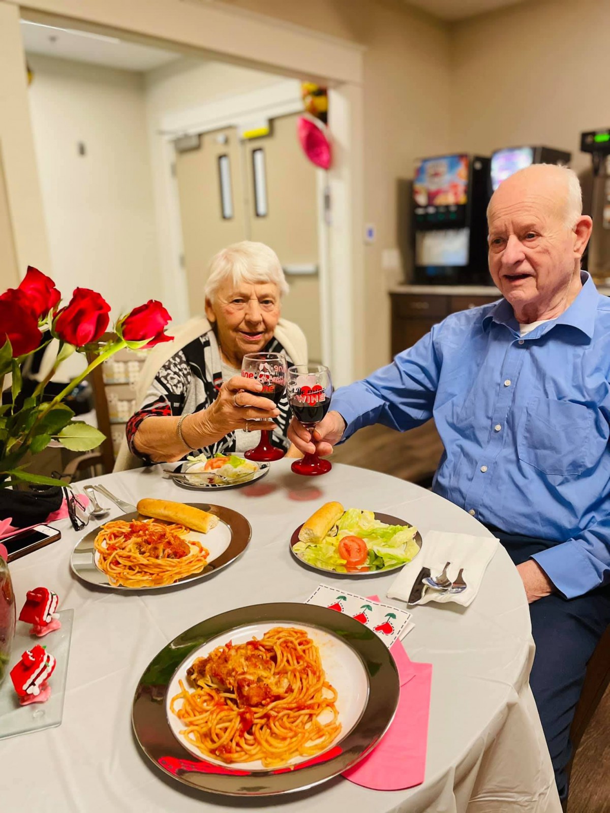An elderly couple enjoys a meal together, featuring spaghetti and wine, at a beautifully set table with red roses.