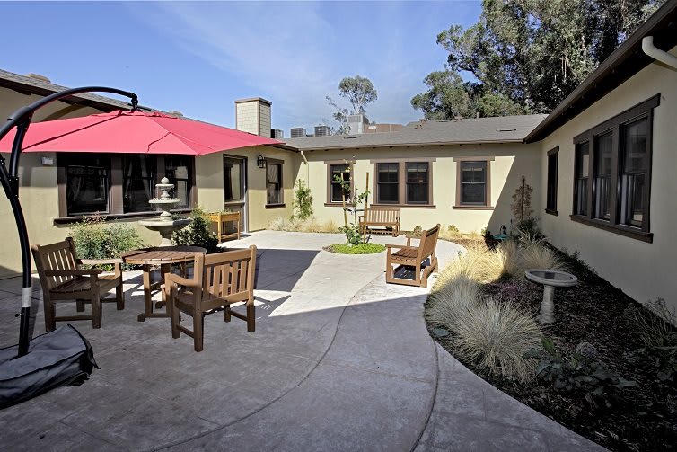 A serene courtyard featuring a patio with wooden furniture, a red umbrella, and a central fountain surrounded by landscaping.