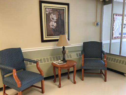A cozy waiting area featuring two blue chairs, a small wooden table with a lamp, and framed artwork on the wall.