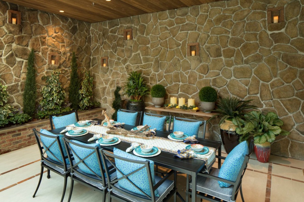An elegant outdoor dining area with a stone wall backdrop, featuring blue cushioned chairs and a well-set table surrounded by lush greenery.