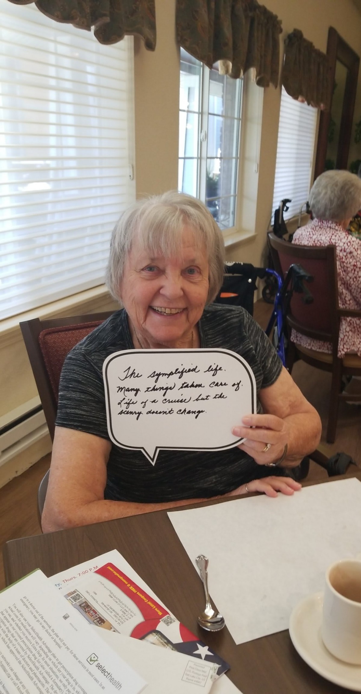 An elderly woman smiling while holding a speech bubble sign in a senior living facility.