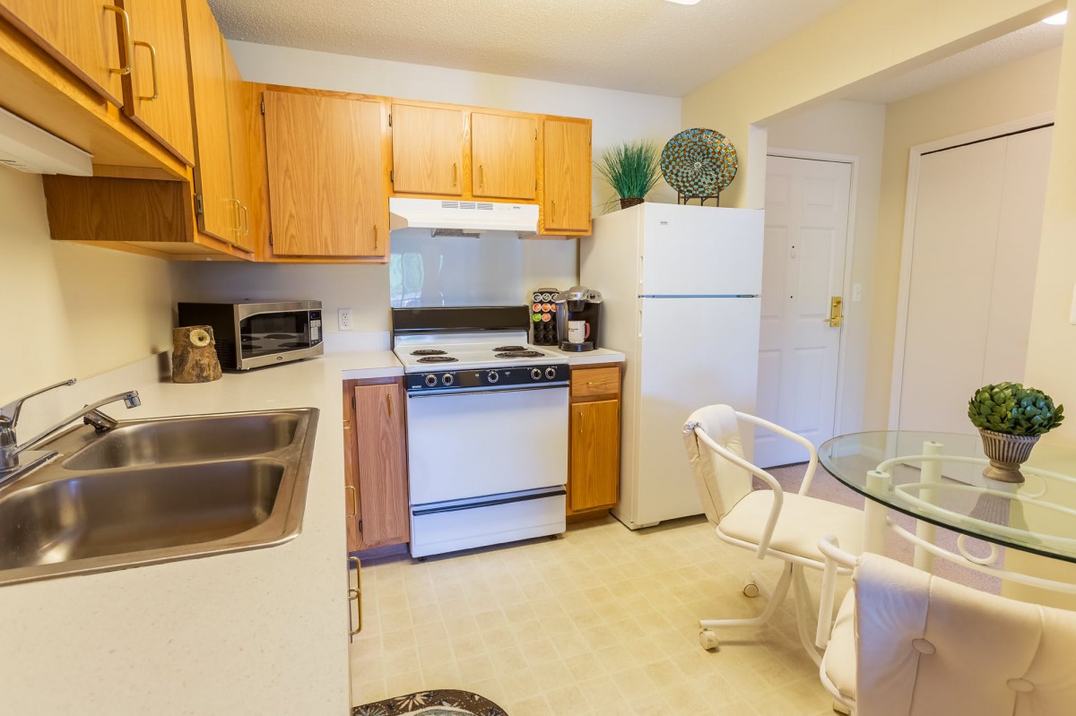 A compact and bright kitchen with wooden cabinets, featuring a refrigerator, oven, and small dining area.