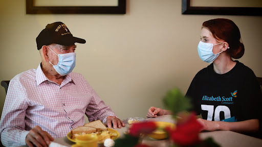 An elderly man and a caregiver having a conversation over a meal while wearing masks.