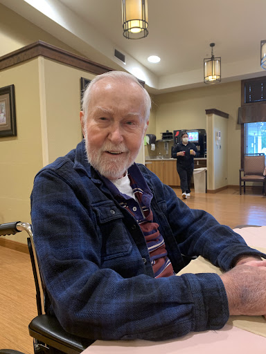 An elderly man sitting at a table in a cozy indoor setting, sharing a smile.