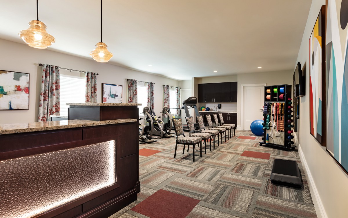 A well-equipped fitness room featuring exercise machines, free weights, and chairs arranged on a patterned carpet.
