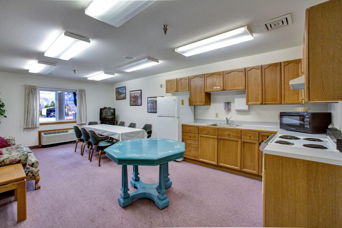 A community room with a kitchen setup, featuring wood cabinets, a refrigerator, tables, and chairs.