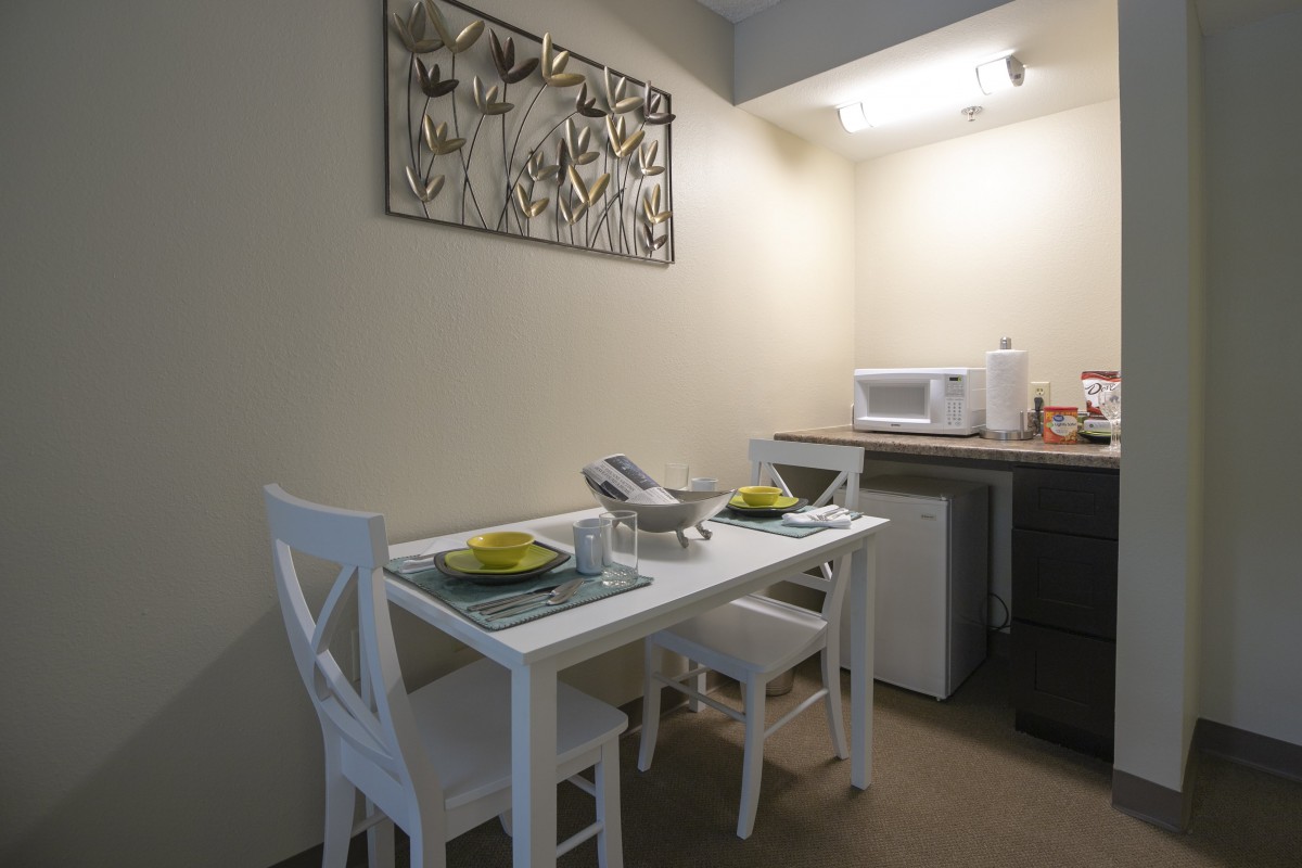 A minimalist dining area with a white table and chairs, set for two, next to a kitchenette.