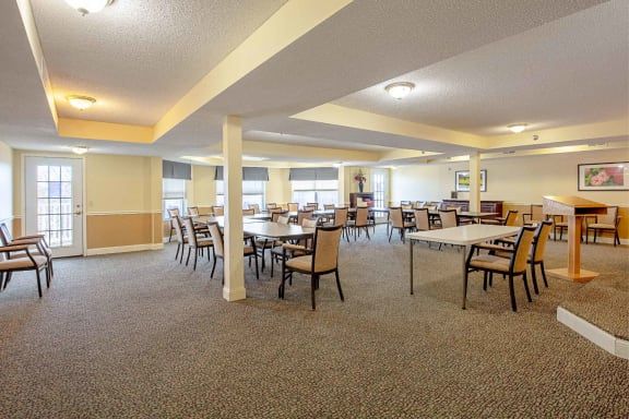 A spacious dining room with multiple tables and chairs, featuring carpeted flooring and ample lighting.