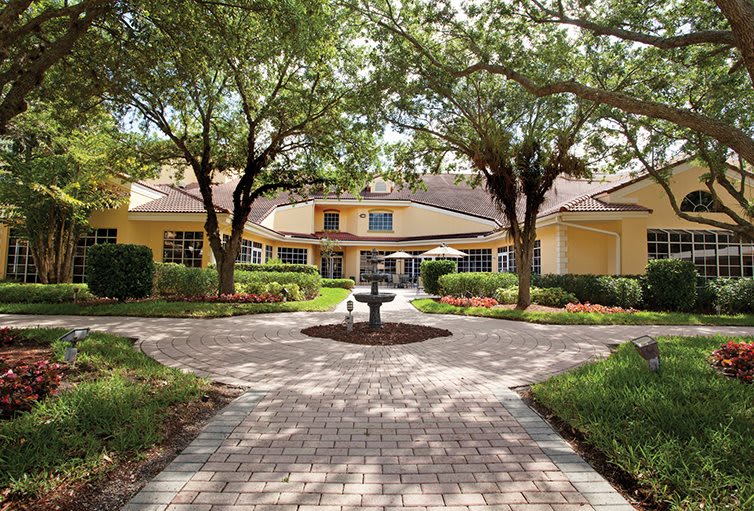 A landscaped courtyard with a fountain and pathway leading to a building surrounded by trees.