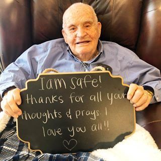 An elderly man smiling while holding a sign that reads 'I am safe! Thanks for all your thoughts & prayers. I love you all!'