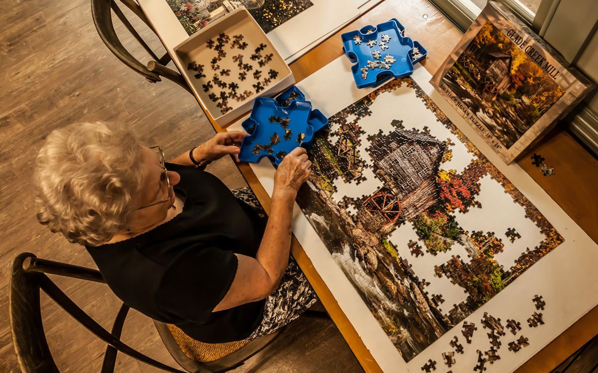 An elderly person assembling a complex jigsaw puzzle on a table, with several puzzle pieces organized in trays.