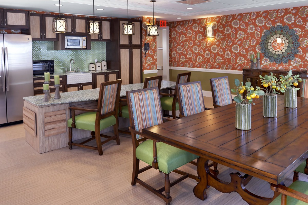 A cozy dining area with a large wooden table, cushioned chairs, and warm lighting, featuring floral wallpaper in the background.