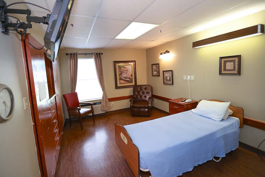 A well-lit hospital room featuring a single bed, a comfortable chair, and framed artwork.