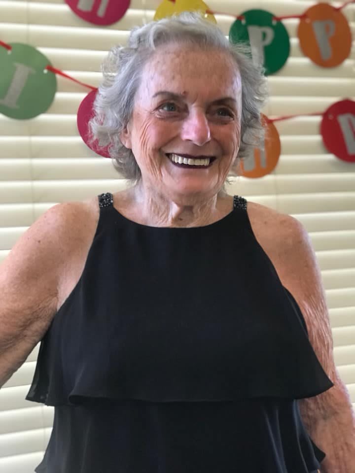 An elderly woman smiling brightly at a celebration, wearing a black dress.