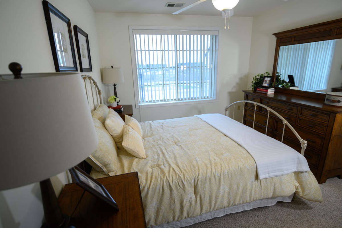 A cozy bedroom with a neatly made bed, dresser, and a large window letting in natural light.