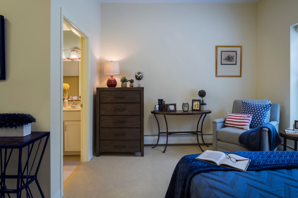 A cozy bedroom with a dresser, table, and comfortable chair, decorated with lamps and framed pictures.