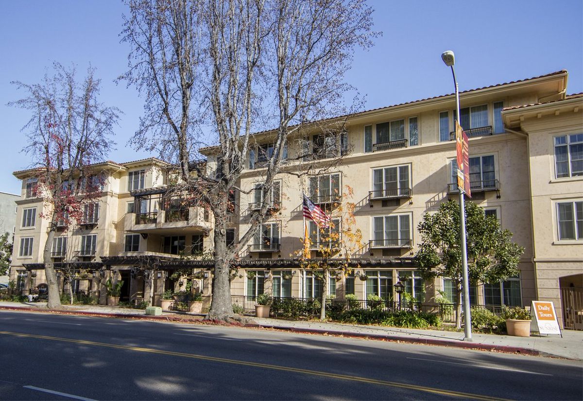 A street view of a multi-story residential building with trees and clear skies.
