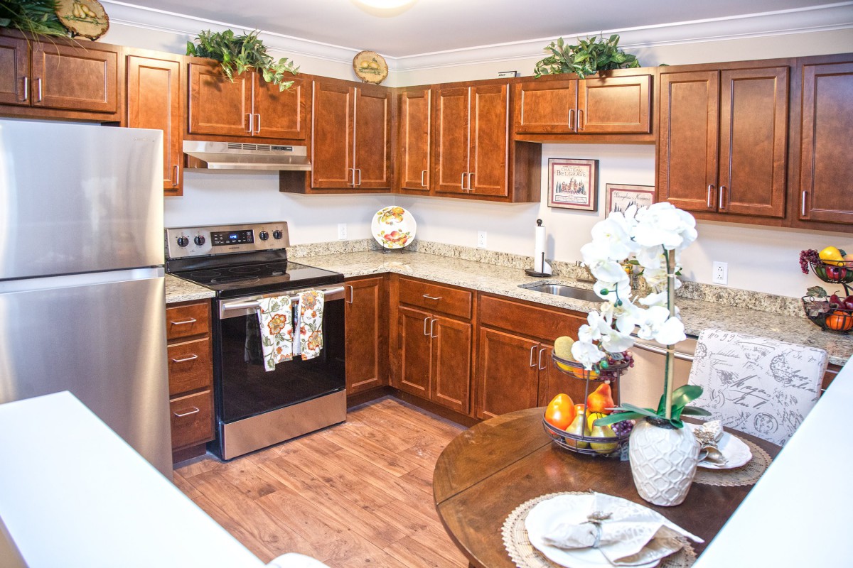 A modern kitchen with wooden cabinets and stainless steel appliances, featuring a corner layout and a small dining area adorned with plants and decorative items.