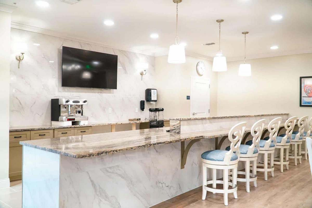 A modern kitchen area featuring a marble countertop with bar stools, a wall-mounted television, and a coffee machine.