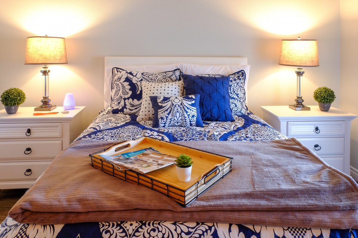 A cozy bedroom setup featuring a neatly arranged bed with patterned pillows, a tray on the blanket, and warm lighting from bedside lamps.