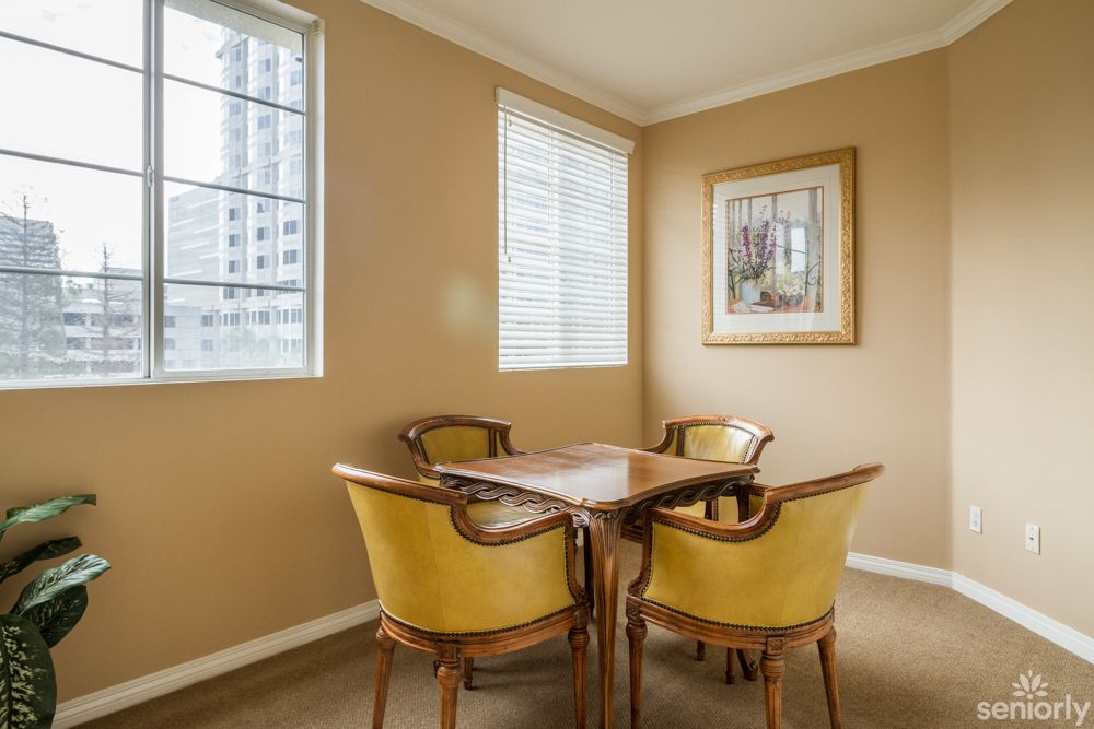 A cozy dining area with a wooden table and four chairs, featuring large windows and a framed painting on the wall.
