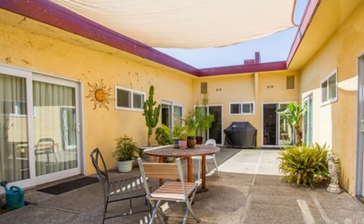 A sunny courtyard featuring a patio with outdoor furniture, surrounded by vibrant yellow walls and various potted plants.