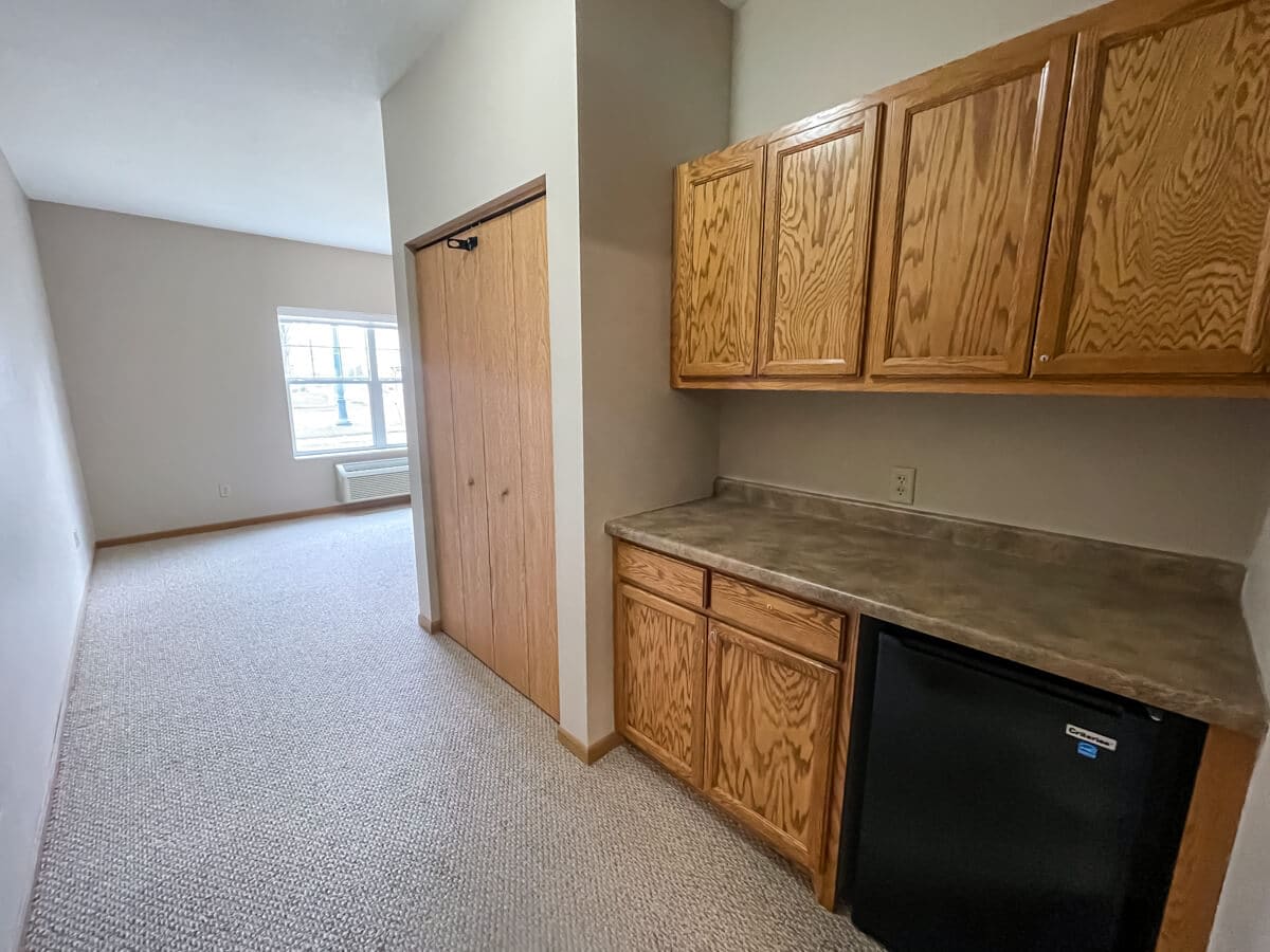 A small kitchenette area in an apartment, featuring wooden cabinets, a countertop, and a mini refrigerator.