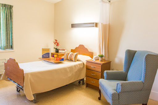 A cozy and well-lit hospital or care facility room featuring a bed, an armchair, and a nightstand.