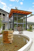 A modern architectural building featuring large glass windows, a wooden roof overhang, and a stone water feature in a courtyard.