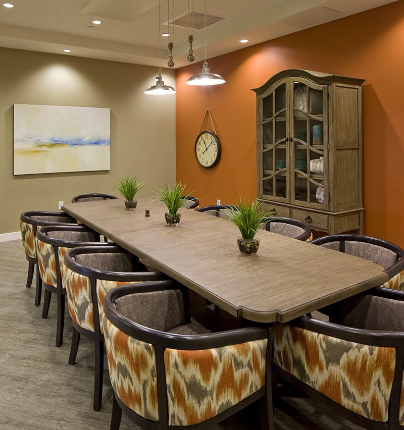 A modern conference room with a large wooden table, surrounded by upholstered chairs. The room is decorated with plants, artwork, and warm lighting.
