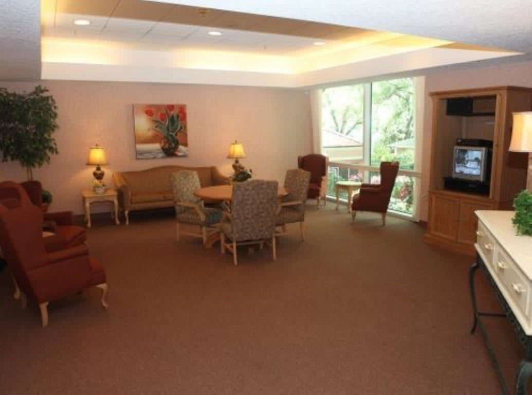 A cozy living room with a seating area featuring a table, several chairs, sofas, and lamps, complemented by a large window and decorative elements.